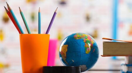 A imagem mostra uma mesa escolar, contendo um porta-canetas laranja com lápis azul e roxo, um globo e livros; e ilustra o artigo sobre Lei de Diretrizes e Bases da Educação Nacional (LDB).