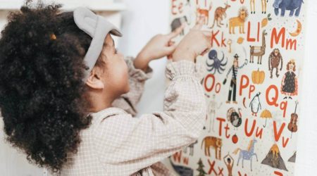 A imagem mostra uma menina apontando as letras do alfabeto em uma parede.