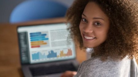 A imagem mostra uma mulher negra sentada em frente ao computador, que mostra gráficos na tela. Utilizamos a imagem para ilustrar nosso artigo sobre as funções de coordenador(a) do CRAS e CREAS.