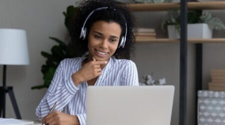 A imagem mostra uma mulher negra olhando para a tela do computador. Ela utiliza fones de ouvido e há uma estante no ambiente. A imagem ilustra o artigo sobre cursos para assistentes sociais.
