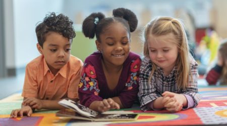 A imagem mostra três crianças em um tapete colorido lendo um livro e ilustra o artigo sobre livros sobre diversidade para ter na biblioteca da escola.